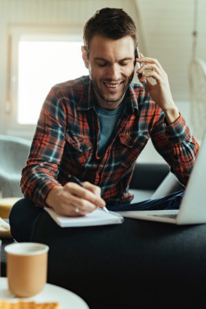 Happy man taking notes while talking on the phone at home.